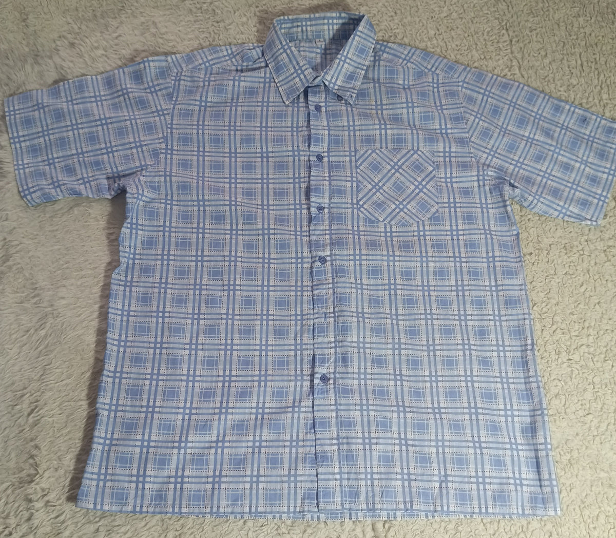 A short-sleeve button-up shirt with a blue and white grid pattern, laid flat on a beige shag rug.