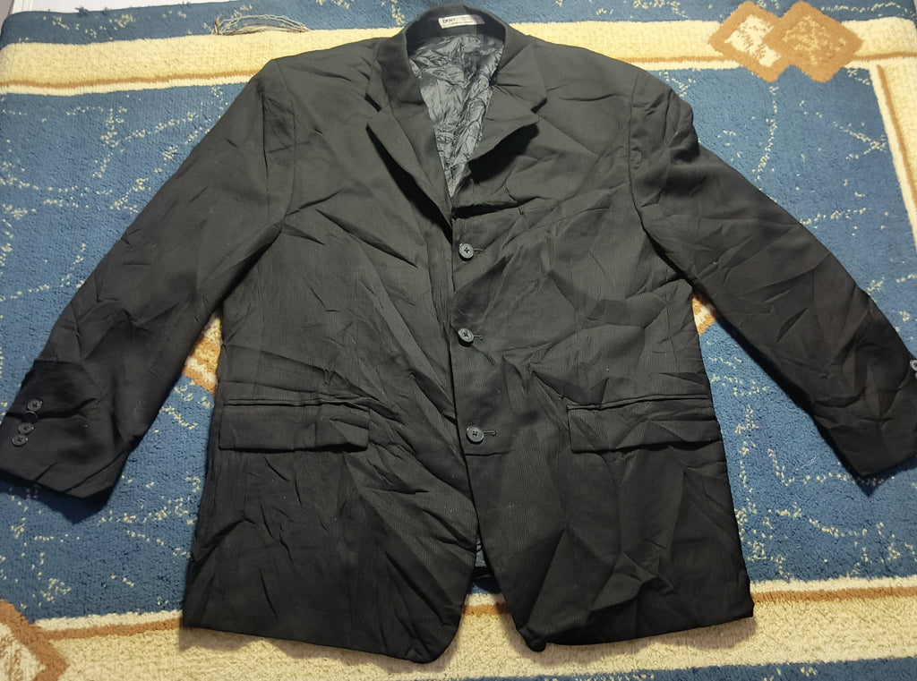A black crinkled jacket laid flat on a blue and gold patterned blanket on a white table, with a gray stool underneath and a camera tripod leg visible in the background.