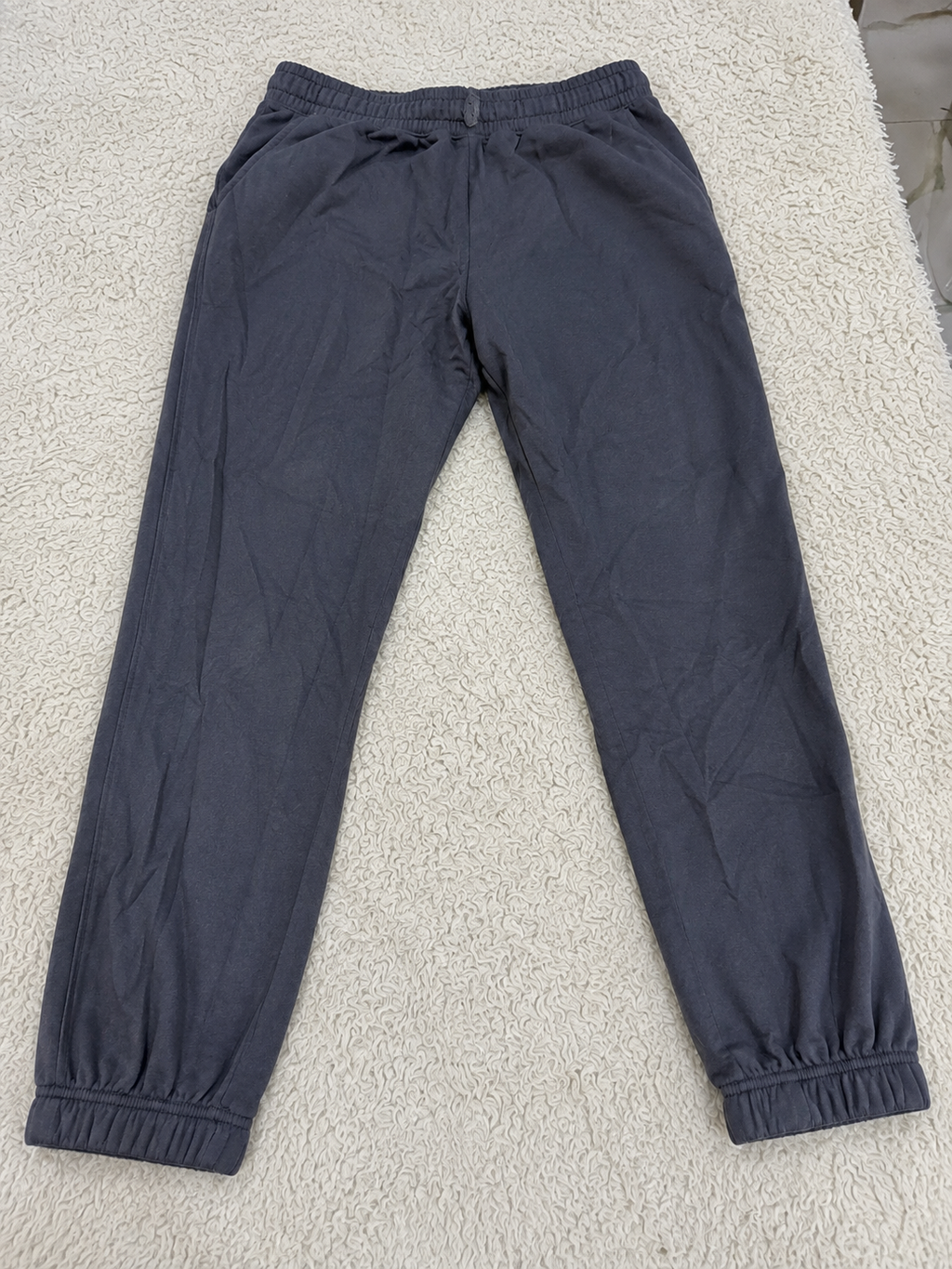 Dark gray jogger pants with an elastic waistband and elastic ankle cuffs, laid flat on a white textured rug.