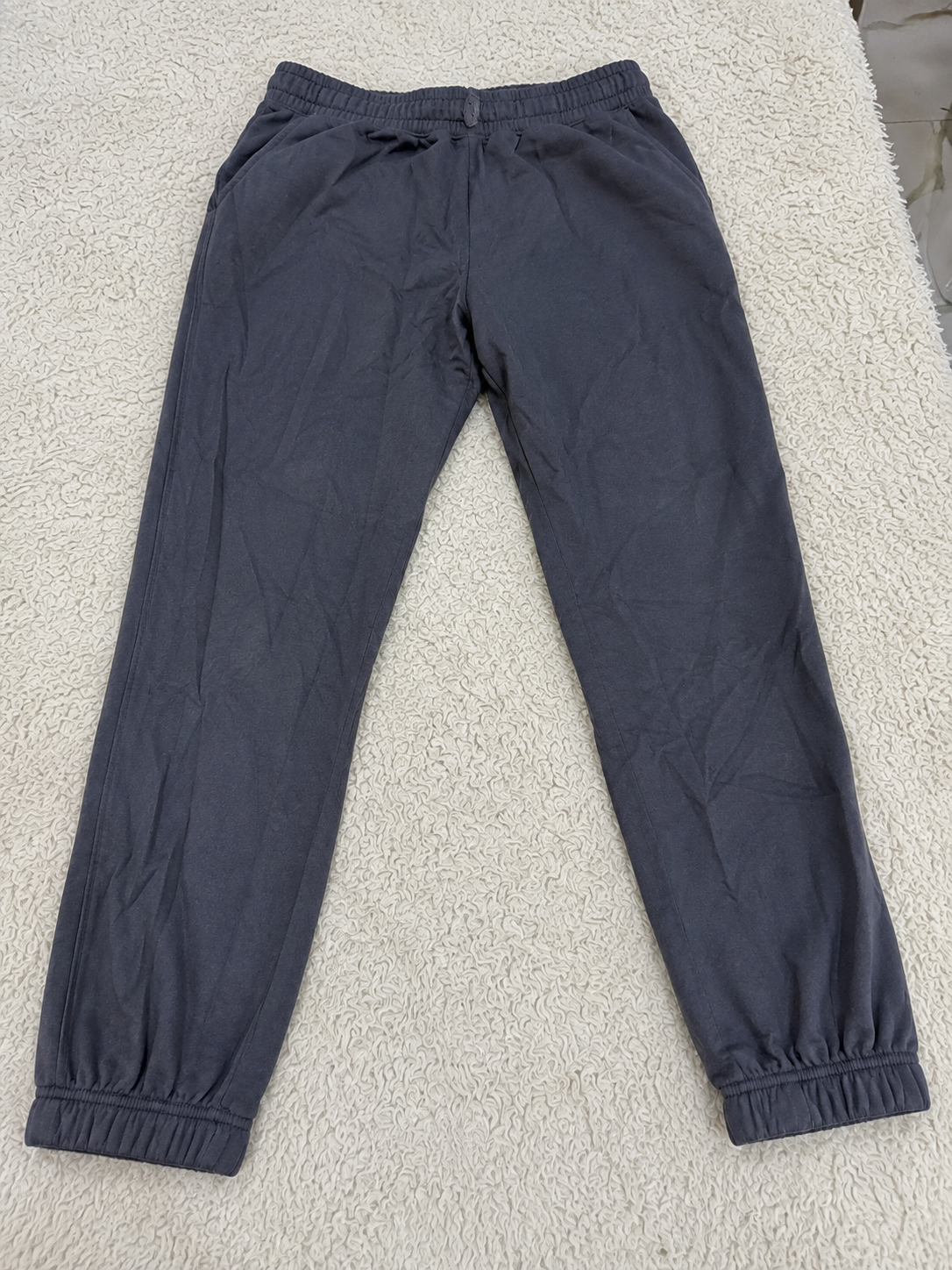 Dark gray jogger pants with an elastic waistband and elastic ankle cuffs, laid flat on a white textured rug.