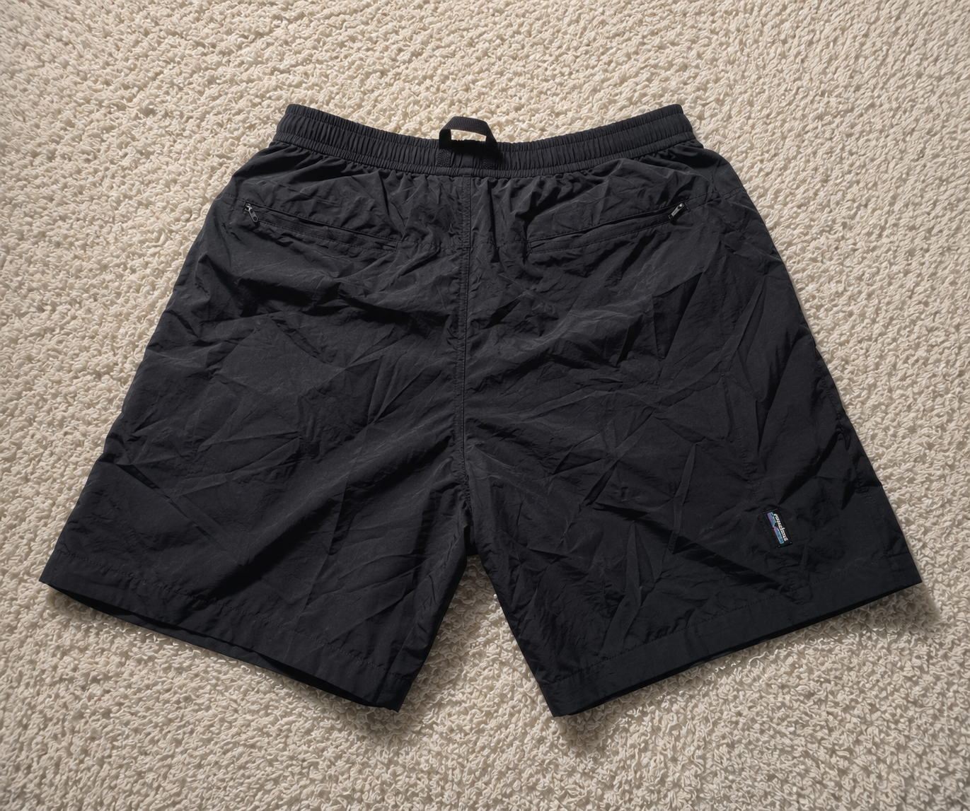 Black lightweight shorts with an elastic waistband and zippered side pockets, laid flat on a beige carpet background.